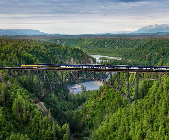 A bridge spans a river surrounded by lush green hills and dense forest in Alaska.