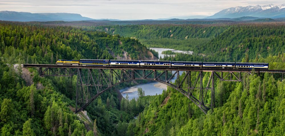 A bridge spans a river surrounded by lush green hills and dense forest in Alaska.