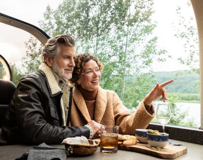 A couple enjoys a scenic train ride, sharing snacks and drinks while admiring the lush landscapes through panoramic windows.