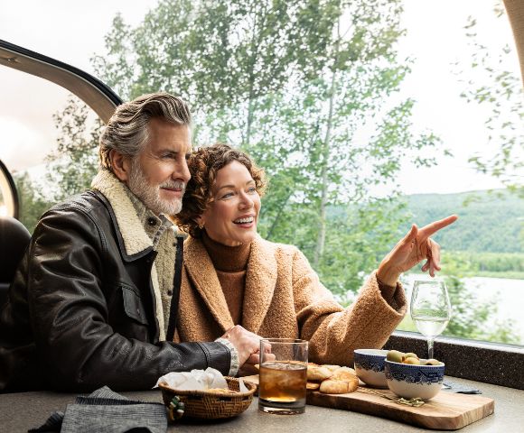 A couple enjoys a scenic train ride, sharing snacks and drinks while admiring the lush landscapes through panoramic windows.