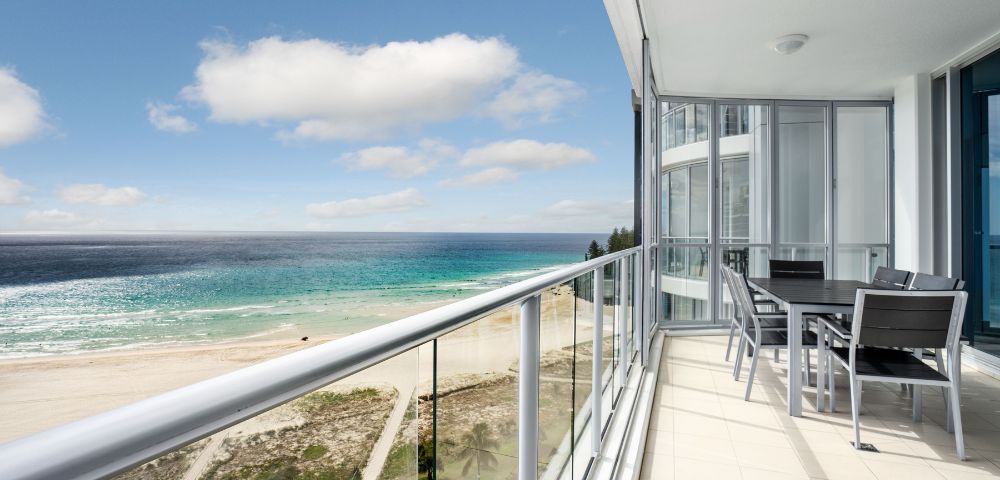 Balcony view with a glass railing overlooking the ocean and beach, featuring outdoor seating.
