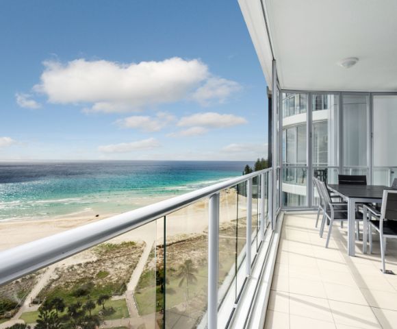 Balcony view with a glass railing overlooking the ocean and beach, featuring outdoor seating.