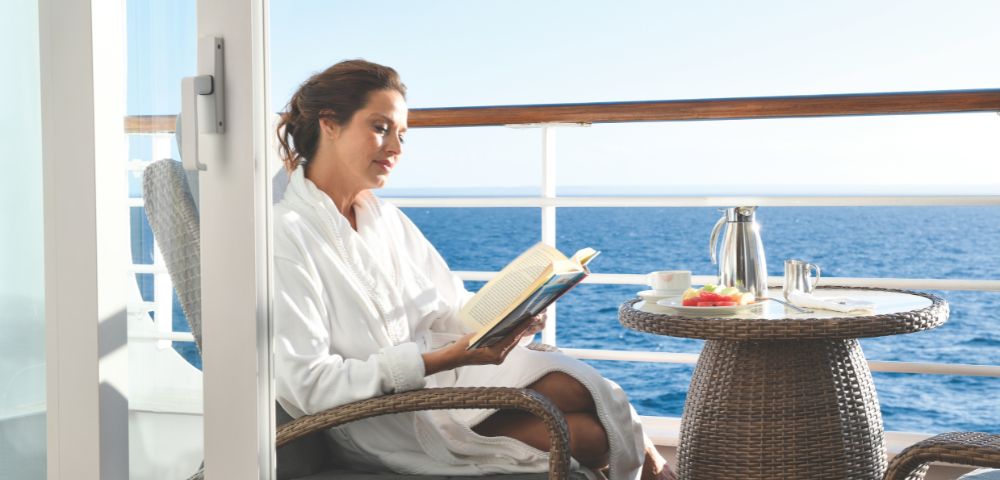 A person in a soft white robe relaxes on a balcony, reading a book with an ocean view and a small table set with refreshments.