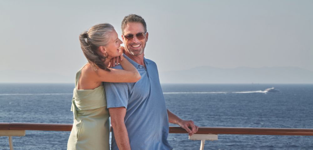 A couple stands together on a cruise ship deck, gazing at the ocean, with mountains just visible on the horizon.