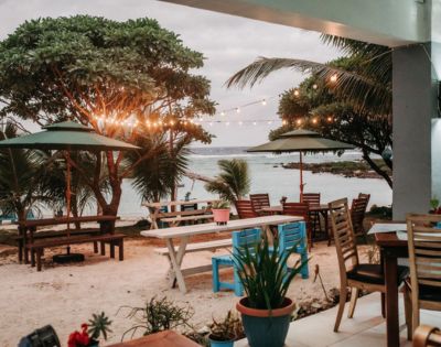 Outdoor dining area with tables and chairs near palm trees, overlooking the water with warm lighting for a cozy seaside meal.