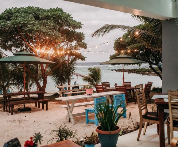 Outdoor dining area with tables and chairs near palm trees, overlooking the water with warm lighting for a cozy seaside meal.