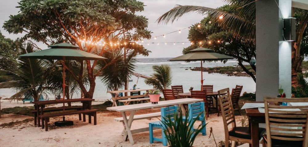 Outdoor dining area with tables and chairs near palm trees, overlooking the water with warm lighting for a cozy seaside meal.