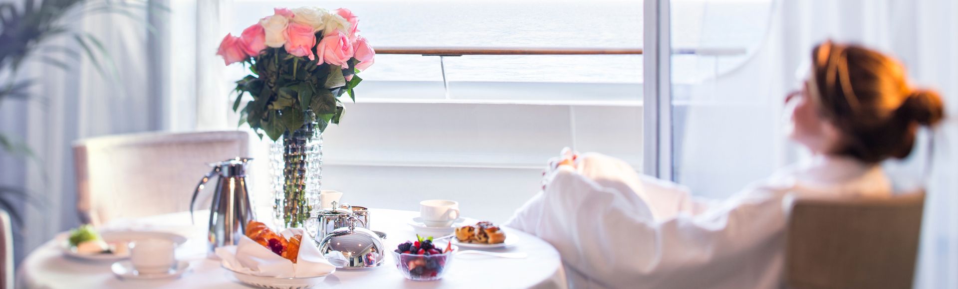 A person relaxes in a robe beside a table set with breakfast and flowers, enjoying ocean views through a balcony window.