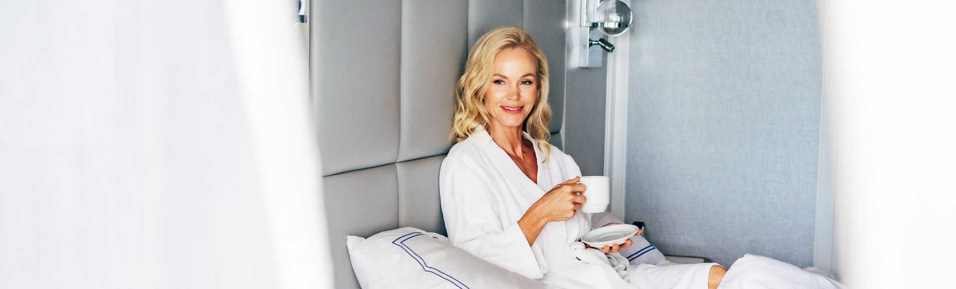 A person in a white robe sips a drink while lounging on a neatly made bed with soft, white linens in a modern, stateroom backdrop onboard Oceania Regatta.