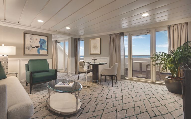 Stylish cruise stateroom featuring a cozy sitting area, dining table, large windows onto balcony with ocean views, and modern decor.