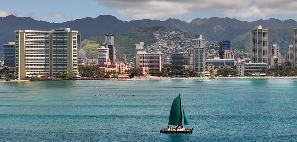 Scenic coastal city view with tall buildings and mountains in the background, featuring a green-sailed sailboat on the water and a beach along the shoreline.