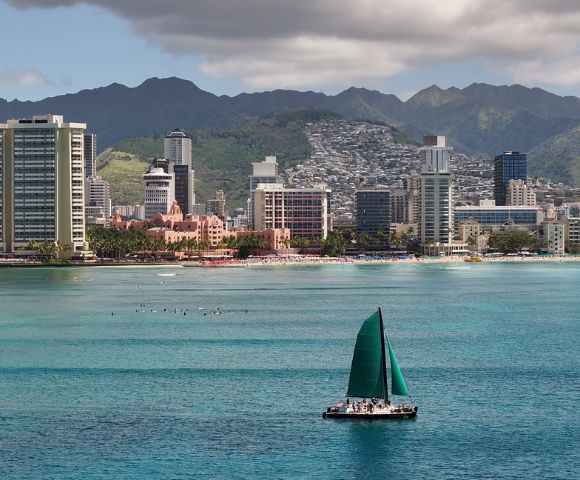 Scenic coastal city view with tall buildings and mountains in the background, featuring a green-sailed sailboat on the water and a beach along the shoreline.