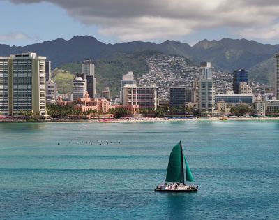 Scenic coastal city view with tall buildings and mountains in the background, featuring a green-sailed sailboat on the water and a beach along the shoreline.
