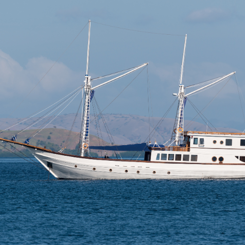 A traditional sailing ship with two masts glides across calm blue waters, surrounded by rolling hills and a clear sky.