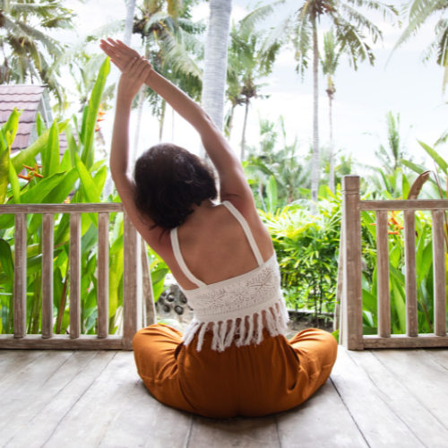 The image shows a woman sitting cross-legged on a wooden deck, facing away from the camera, with her arms raised above her head in a stretching or yoga pose. She is surrounded by lush, tropical greenery, including tall palm trees and broad-leaved plants, creating a serene and natural backdrop. The setting appears to be a tropical or island location, emphasizing relaxation and tranquility amidst the vibrant, green environment.