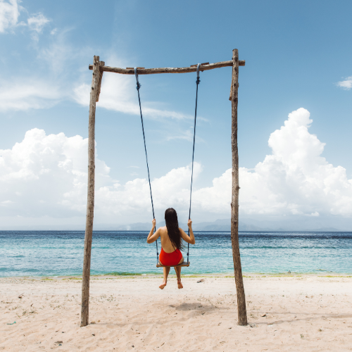 The image depicts a girl sitting on a swing on a sandy beach, facing the ocean. She is wearing a red dress and has long dark hair flowing down her back. The swing is supported by a simple wooden frame made of roughly hewn logs, and the sky above is bright with scattered clouds. The calm blue sea stretches out to the horizon, creating a peaceful and idyllic seaside scene.