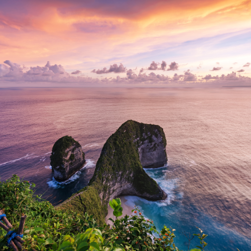 This breathtaking image captures a stunning coastal scene during sunset, with a sky painted in warm hues of orange, pink, and purple. In the foreground, lush green foliage and blooming plants frame the view of dramatic limestone cliffs rising from the ocean. These towering rock formations are partially covered in greenery and surrounded by gentle waves crashing against their bases. The tranquil sea extends to the horizon, creating a harmonious blend of vibrant sky and serene water, evoking a sense of peaceful natural beauty.