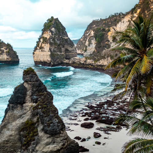 The image depicts a stunning coastal scene featuring towering limestone cliffs and rock formations rising dramatically from the turquoise ocean. A sandy beach with scattered rocks curves along the shoreline, bordered by lush green palm trees on the right side. The sky above is partly cloudy, adding a touch of moody atmosphere to the beautiful tropical landscape.