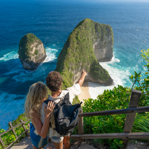 The image shows two travelers, a man and a woman, standing on a wooden staircase overlooking a stunning coastal landscape. The man, pointing towards the horizon, has curly hair and is wearing a backpack, while the woman, with long blonde hair, is holding onto his shoulder. They are surrounded by lush green foliage, and in the background, dramatic limestone cliffs rise from the turquoise ocean, with waves crashing against their bases. The scene captures a moment of awe and adventure amid breathtaking natural beauty.