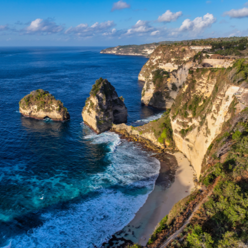 The image captures a stunning coastal landscape featuring dramatic white cliffs and rock formations rising sharply from the deep blue ocean. A small sandy beach nestled at the base of the cliffs is visible, with waves gently crashing onto the shore. The lush greenery atop the cliffs contrasts beautifully with the rugged rocks and vibrant sea, creating a breathtaking scene of natural beauty and rugged coastline.