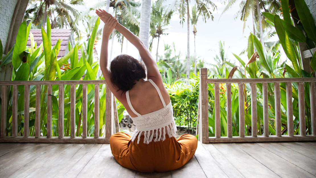 The image shows a woman sitting cross-legged on a wooden deck, facing away from the camera, with her arms raised above her head in a stretching or yoga pose. She is surrounded by lush, tropical greenery, including tall palm trees and broad-leaved plants, creating a serene and natural backdrop. The setting appears to be a tropical or island location, emphasizing relaxation and tranquility amidst the vibrant, green environment.
