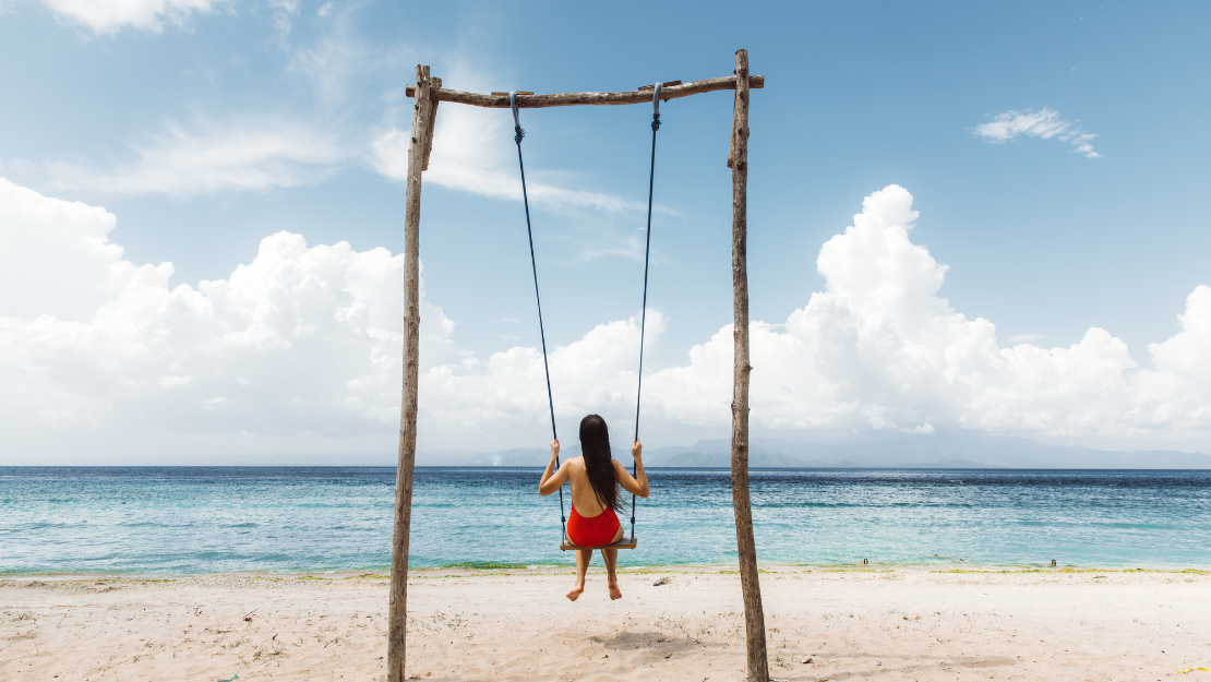The image depicts a girl sitting on a swing on a sandy beach, facing the ocean. She is wearing a red dress and has long dark hair flowing down her back. The swing is supported by a simple wooden frame made of roughly hewn logs, and the sky above is bright with scattered clouds. The calm blue sea stretches out to the horizon, creating a peaceful and idyllic seaside scene.