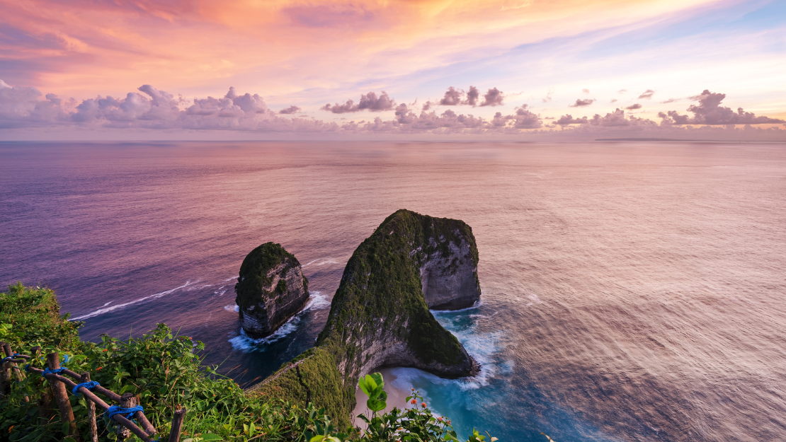 This breathtaking image captures a stunning coastal scene during sunset, with a sky painted in warm hues of orange, pink, and purple. In the foreground, lush green foliage and blooming plants frame the view of dramatic limestone cliffs rising from the ocean. These towering rock formations are partially covered in greenery and surrounded by gentle waves crashing against their bases. The tranquil sea extends to the horizon, creating a harmonious blend of vibrant sky and serene water, evoking a sense of peaceful natural beauty.