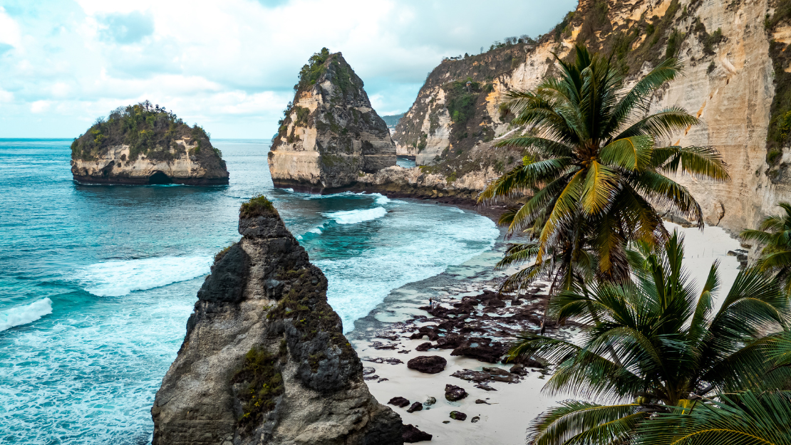 The image depicts a stunning coastal scene featuring towering limestone cliffs and rock formations rising dramatically from the turquoise ocean. A sandy beach with scattered rocks curves along the shoreline, bordered by lush green palm trees on the right side. The sky above is partly cloudy, adding a touch of moody atmosphere to the beautiful tropical landscape.