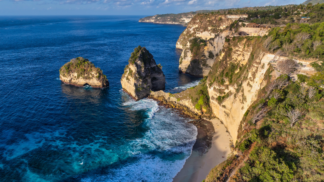 The image captures a stunning coastal landscape featuring dramatic white cliffs and rock formations rising sharply from the deep blue ocean. A small sandy beach nestled at the base of the cliffs is visible, with waves gently crashing onto the shore. The lush greenery atop the cliffs contrasts beautifully with the rugged rocks and vibrant sea, creating a breathtaking scene of natural beauty and rugged coastline.
