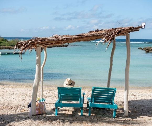 Idyllic beach scene with clear blue water, white sand, and lounge chairs under a wooden canopy.