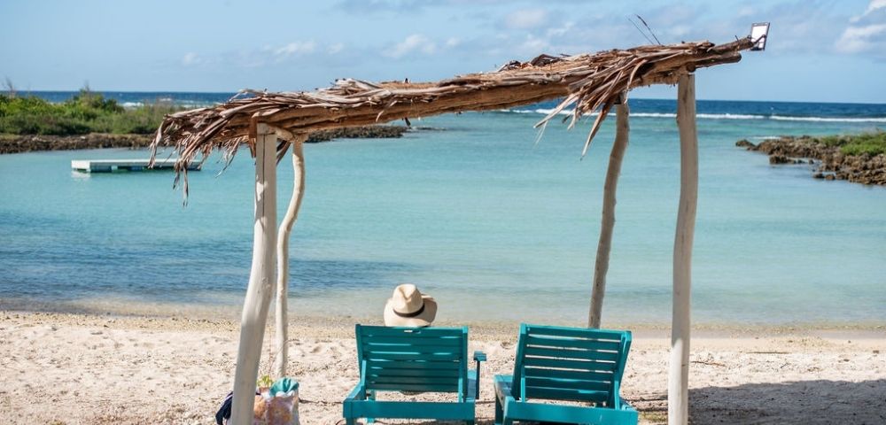 Idyllic beach scene with clear blue water, white sand, and lounge chairs under a wooden canopy.