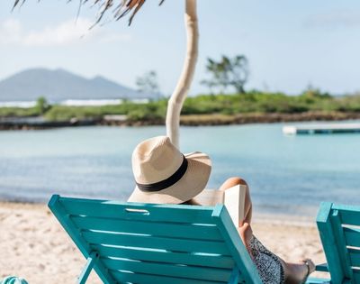 Person sitting on a lounge chair looking at the ocean, wearing a hat.
