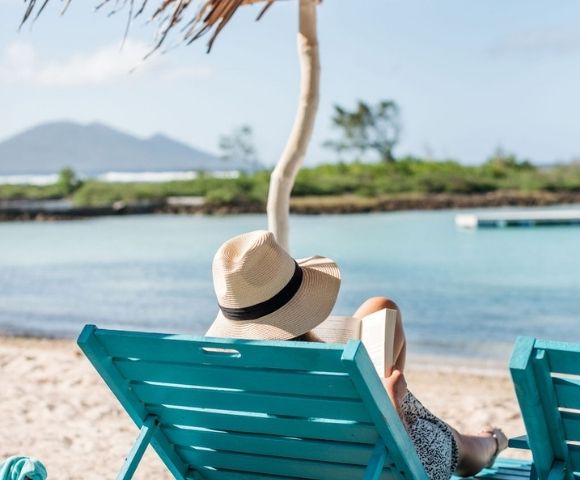 Person sitting on a lounge chair looking at the ocean, wearing a hat.