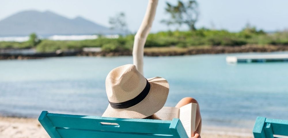 Person sitting on a lounge chair looking at the ocean, wearing a hat.