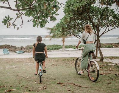 Person riding a bicycle along a shoreline path near a water body under cloudy skies.