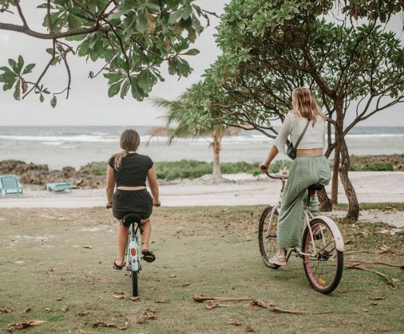 Person riding a bicycle along a shoreline path near a water body under cloudy skies.