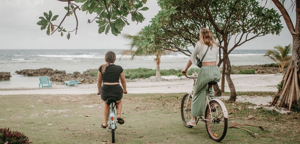 Person riding a bicycle along a shoreline path near a water body under cloudy skies.