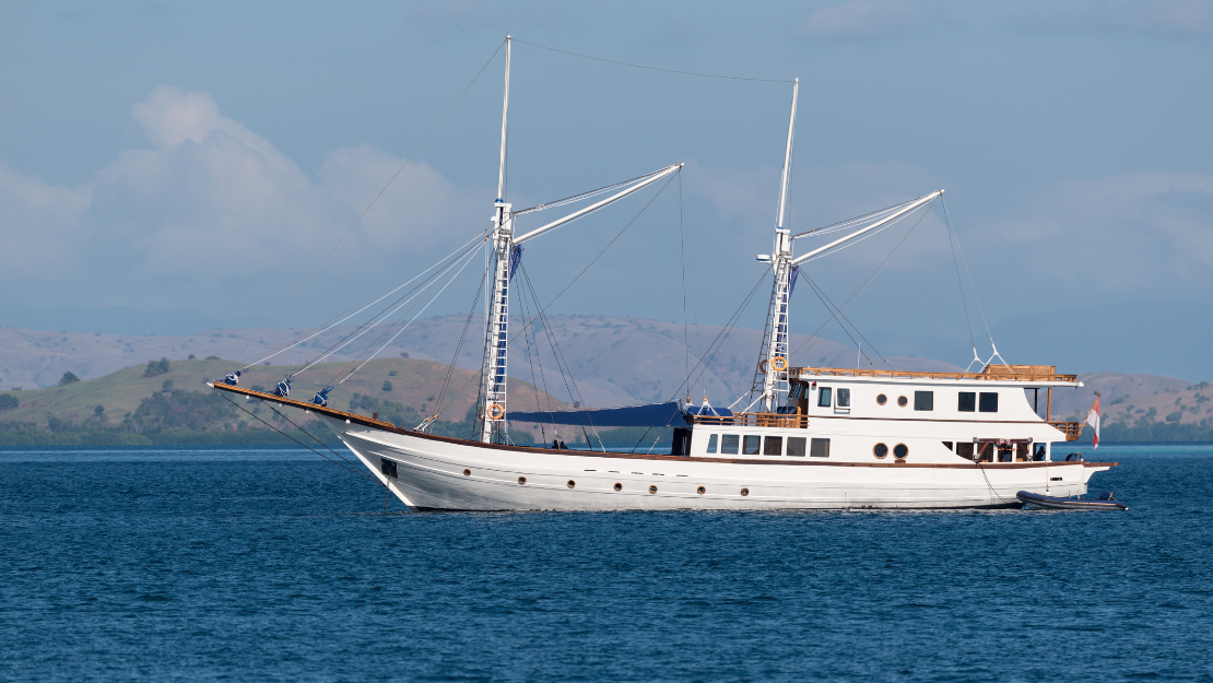 A traditional sailing ship with two masts glides across calm blue waters, surrounded by rolling hills and a clear sky.