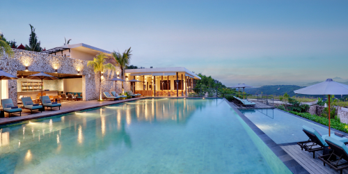 Luxurious resort with a tranquil infinity pool, palm trees, and modern architecture against a scenic hillside backdrop at dusk.