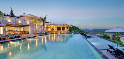 Luxurious resort with a tranquil infinity pool, palm trees, and modern architecture against a scenic hillside backdrop at dusk.