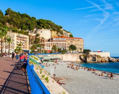 Scenic coastal area with a beach and promenade, featuring people sunbathing, palm trees, colorful buildings, and green hills in the background under a clear sky with wispy clouds.