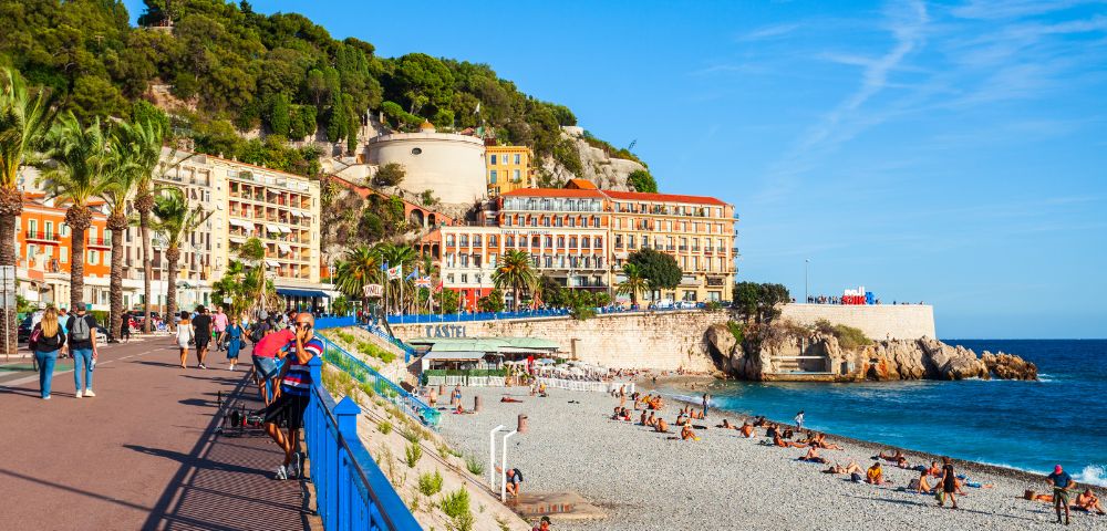Scenic coastal area with a beach and promenade, featuring people sunbathing, palm trees, colorful buildings, and green hills in the background under a clear sky with wispy clouds.