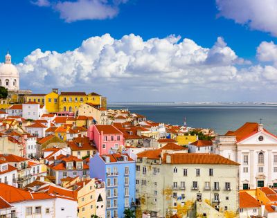Colorful cityscape of Lisbon, Portugal, showcasing red-tiled rooftops, pastel-colored buildings, a white dome, a church with a bell tower, and the Tagus River under a partly cloudy sky.