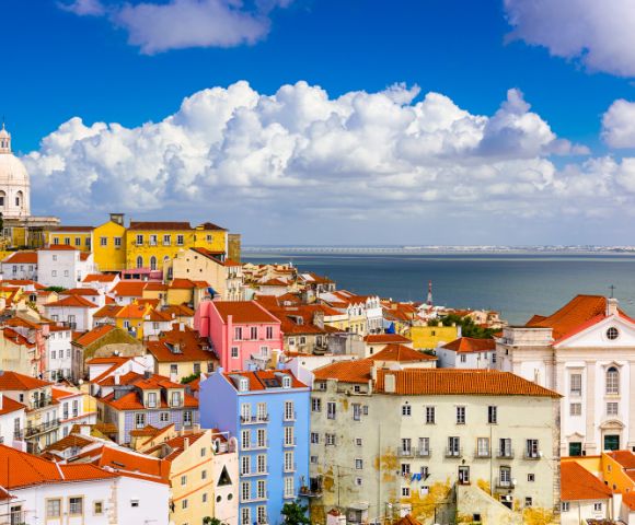 Colorful cityscape of Lisbon, Portugal, showcasing red-tiled rooftops, pastel-colored buildings, a white dome, a church with a bell tower, and the Tagus River under a partly cloudy sky.
