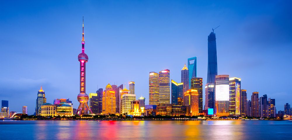 Shanghai skyline at dusk, featuring illuminated skyscrapers reflected in the calm river. The scene conveys a vibrant, futuristic cityscape.