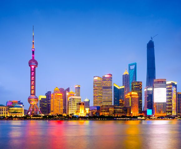 Shanghai skyline at dusk, featuring illuminated skyscrapers reflected in the calm river. The scene conveys a vibrant, futuristic cityscape.