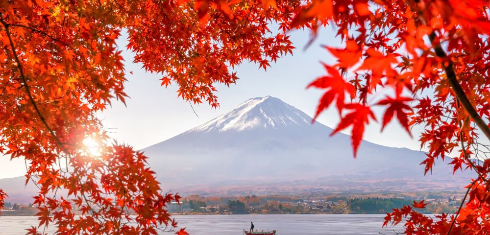 A person stands on a boat in a lake beneath snow-capped Mount Fuji, framed by vivid red autumn leaves.