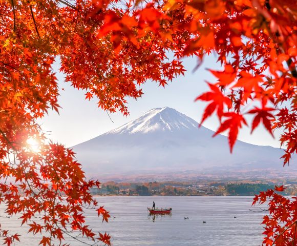 A person stands on a boat in a lake beneath snow-capped Mount Fuji, framed by vivid red autumn leaves.