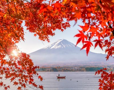 A person stands on a boat in a lake beneath snow-capped Mount Fuji, framed by vivid red autumn leaves.