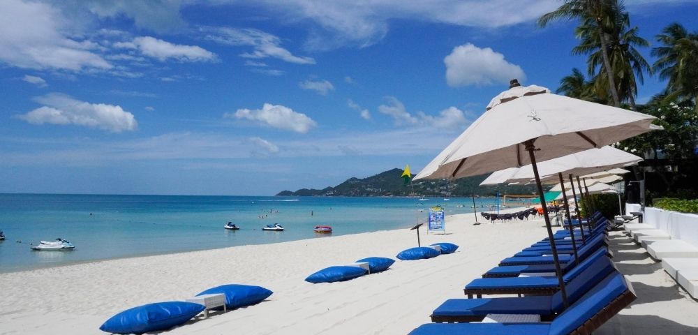 Sunny beach scene with a row of blue lounge chairs and white umbrellas on white sand. Calm turquoise sea, blue sky, a few clouds, and distant hills. Relaxing vibe.